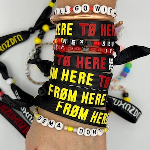 May include: A collection of black fabric wristbands featuring yellow and red text that reads "FROM HERE TO HERE" and "TØ HERE", alongside beaded bracelets with white letter beads spelling "I'LL GO WITH", "NEXT", and "DEMA DONT" with yellow and red accents.