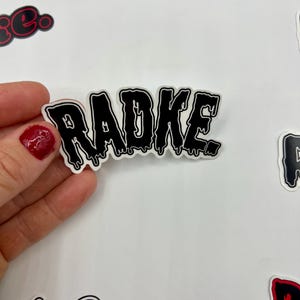 May include: Black and white sticker with the word "RADKE" in dripping letters. The sticker is on a white background.