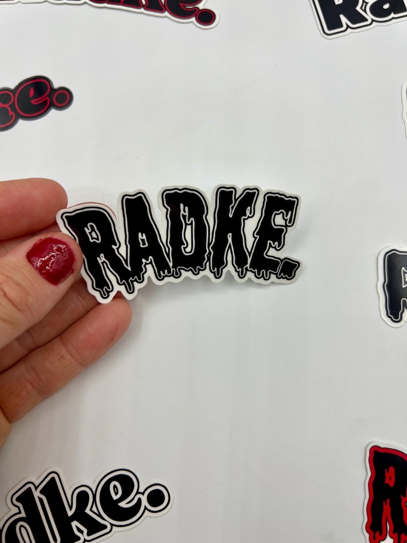 May include: Black and white sticker with the word "RADKE" in dripping letters. The sticker is on a white background.