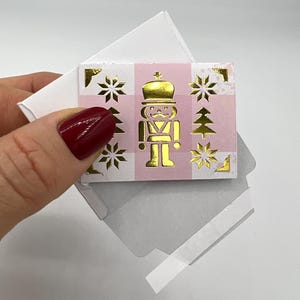 Könnte beinhalten: Kleine quadratische Karte mit einem goldenen Nussknacker-Soldaten-Design auf einem rosa-weiß karierten Hintergrund. Goldene Schneeflocken und Weihnachtsbäume verzieren die Ränder. Die Karte wird in einem weißen Umschlag gehalten.