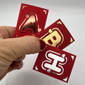 Puede incluir: Tres tarjetas rectangulares rojas con letras A, B y H en lámina metálica. Cada tarjeta tiene una superficie texturizada, detalles decorativos en las esquinas y pequeñas formas de corazón alrededor de la letra central. Las letras son de oro rosa, oro y plata.