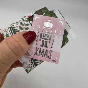 Peut inclure: Un ensemble d'étiquettes cadeaux de Noël. L'étiquette supérieure est rose avec un motif de casse-noisette argenté et le mot "XMAS". D'autres étiquettes présentent des motifs de houx et de feuilles en vert et blanc. Les étiquettes sont maintenues ensemble avec du ruban adhésif blanc.