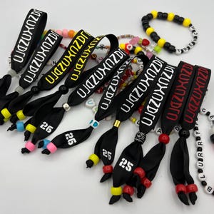 May include: A selection of black wristbands with white text reading "JAZUXOZDRO" and colourful beaded bracelets. Some wristbands have the number "26" printed on them. The bracelets feature various colours and bead sizes.