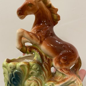 May include: A ceramic horse figurine in shades of brown, tan, and green. The horse is rearing up, with its front legs raised. The base features a decorative, leafy design. The figurine is likely a planter or decorative piece.