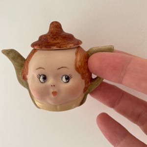 May include: A small, vintage ceramic teapot with a face design. The teapot has a light skin tone, red hair, and blue eyes. The lid and handle are brown and green. The spout and handle are green. A unique decorative item.