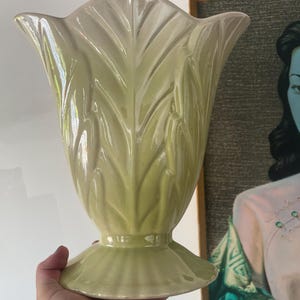 Puede incluir: Un jarrón de cerámica verde pálido con la parte superior acampanada y una base de pedestal. El jarrón tiene un diseño en forma de hoja. El jarrón está sostenido por una mano, con una pintura enmarcada al fondo.