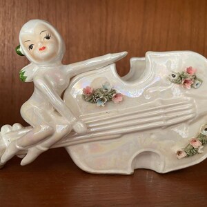 May include: A white ceramic figurine of a fairy playing a white ceramic violin with pink and blue flowers. The fairy has a pink dress and green wings.