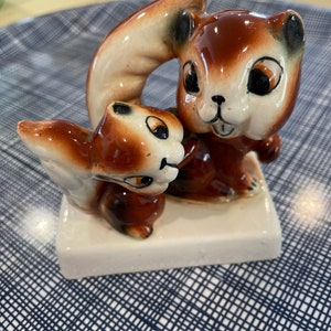 Peut inclure: Une figurine en céramique représentant deux écureuils de dessin animé. Les écureuils sont bruns et blancs avec des détails noirs, posés sur une base rectangulaire blanche. L'un des écureuils tient une grosse noix. L'ornement est un objet décoratif.