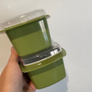 Puede incluir: Dos recipientes de almacenamiento de alimentos verde oliva con tapas de plástico transparente están apilados. Los recipientes tienen forma cuadrada con bordes redondeados y un borde alrededor de la parte superior. Las tapas son transparentes y encajan.