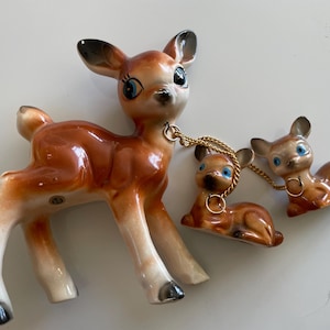 May include: A set of three ceramic deer figurines featuring a large standing fawn and two smaller reclining fawns. The deer are brown with white underbellies, black hooves, and blue eyes. The two smaller fawns are connected to the larger one by gold chains.
