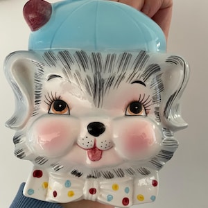 May include: A ceramic dog-shaped cookie jar with a light blue lid and a red accent. The dog has gray fur, pink cheeks, and a polka-dot collar. The jar is held by a person.