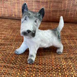 May include: A ceramic Scottish Terrier dog figurine. The dog is white with grey and black markings on its head, ears, and tail. The figurine is posed in a standing position with its tail up. The dog is on a brown woven surface.