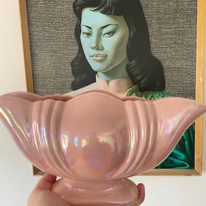 Puede incluir: Una jardinera de cerámica rosa iridiscente con un borde festoneado y una base de pedestal. La jardinera se sostiene frente a un retrato enmarcado de una mujer con cabello negro y tez verde azulado.