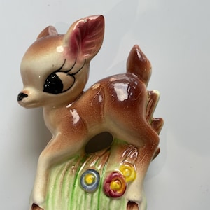 May include: A ceramic fawn figurine with a brown coat, cream face, and large black eyes. The fawn stands on a patch of green grass with colourful flowers. The figurine has a vintage, hand-painted appearance.