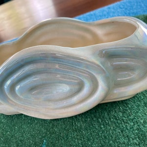 Puede incluir: Una jardinera vintage de forma ovalada con un acabado nacarado. La jardinera de cerámica tiene un color base crema con reflejos iridiscentes azules y verdes. Los lados presentan un diseño arremolinado repetitivo. La jardinera está sobre una superficie verde.