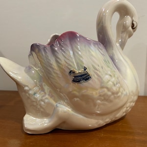 Puede incluir: Un macetero de cerámica blanca en forma de cisne con un esmalte iridiscente. La cabeza del cisne está girada hacia un lado y el macetero tiene un diseño de flores rosas y moradas. El macetero tiene una etiqueta que dice "Olivera Pottery, Pottery St., E. Liverpool, Ohio".