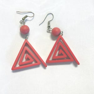 May include: A pair of dangle earrings featuring a red triangular frame with an internal spiral design. Each earring is suspended from a silver-toned metal hook and includes a small, round, matte red bead above the triangular pendant.