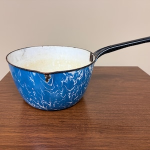 May include: A vintage blue and white enamel saucepan with a black handle. The pan has a marbled pattern and a white interior. The handle is long and straight, and the pan sits on a wooden surface.