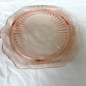 May include: A vintage pink glass serving plate with a scalloped edge and floral detailing. The plate features a central, raised area with radial lines and a decorative border. The plate is likely used for serving cakes or other desserts.