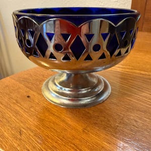 May include: A decorative bowl with a cobalt blue glass interior and a silver-colored metal filigree exterior. The metalwork features geometric cutouts of diamonds, circles, and triangles. The bowl sits on a pedestal base.