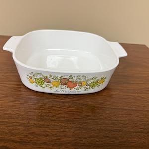 May include: A white square ceramic baking dish with integrated side handles. The exterior features a colorful botanical illustration of various vegetables and herbs, with the cursive text La Marjolaine printed below the design.