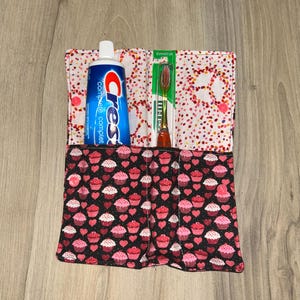Peut inclure: Un organiseur de voyage en tissu avec un motif de cupcakes et de cœurs noirs. Il contient un tube de dentifrice Crest et une brosse à dents. La doublure intérieure présente un motif à pois roses et blancs.
