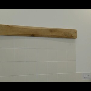 May include: A light wood shelf with a slightly curved shape, mounted on a white tiled wall. The shelf has two small wooden pegs on each end.