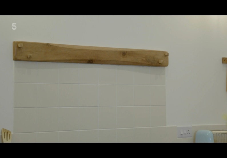 May include: A light wood shelf with a slightly curved shape, mounted on a white tiled wall. The shelf has two small wooden pegs on each end.