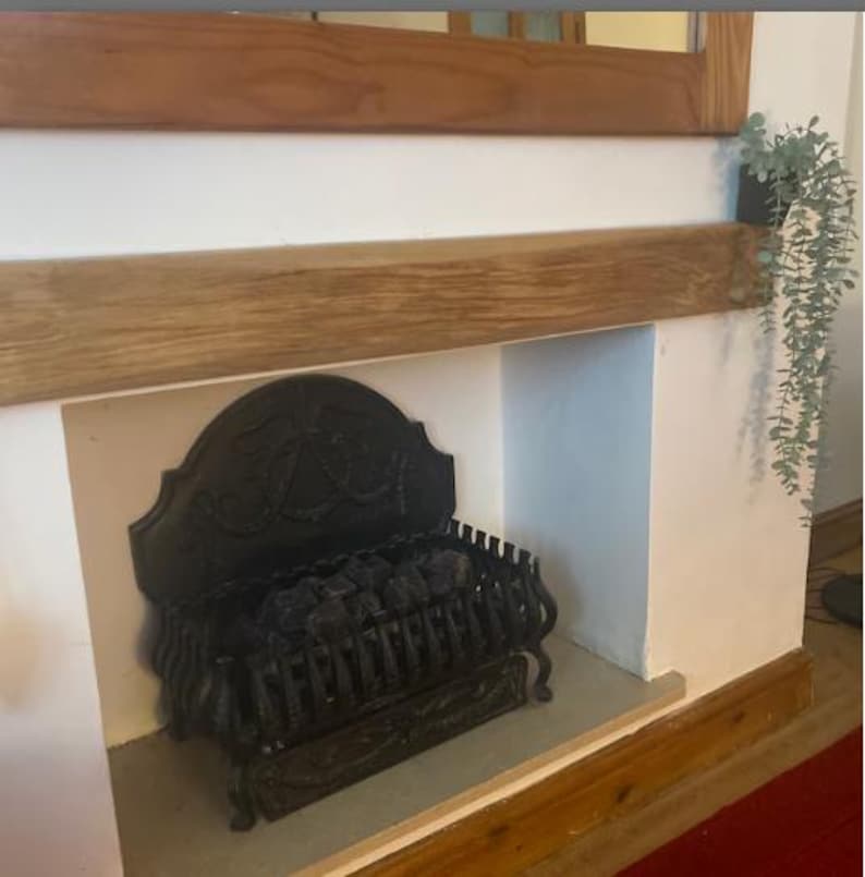 May include: A black cast iron fireplace insert with a decorative design and a grate for burning logs. The fireplace is set into a white wall with a wooden mantel above it.