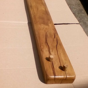 May include: A long, wooden shelf with three wooden pegs. The shelf is stained a light brown color and has a natural wood grain.