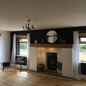 May include: A living room with a fireplace, a wood floor, and white curtains. The fireplace has a black wood-burning stove and a wooden mantel with a round mirror above it. There are two windows with white curtains on either side of the fireplace.