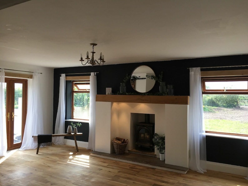 May include: A living room with a fireplace, a wood floor, and white curtains. The fireplace has a black wood-burning stove and a wooden mantel with a round mirror above it. There are two windows with white curtains on either side of the fireplace.