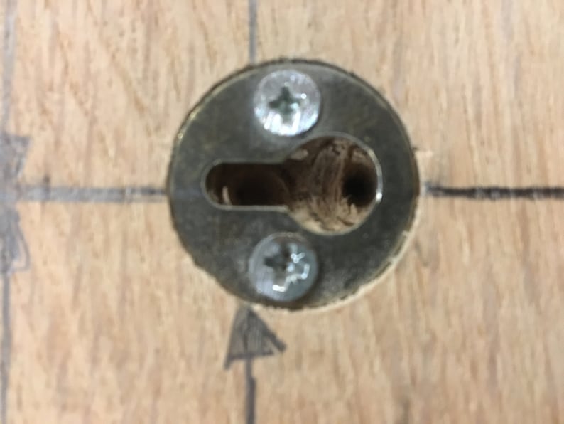 May include: Close-up of a metal keyhole cover with two screws. The cover is mounted on a wooden surface.