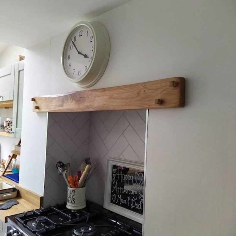 May include: A wooden shelf with a natural finish is mounted above a white tiled backsplash in a kitchen. The shelf has two wooden pegs on each side. A white clock with black hands is mounted on the wall above the shelf. A black framed chalkboard with the text "Happy Anniversary Mum & Dad" is on the counter below the shelf.