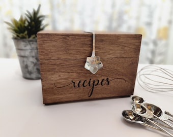 Könnte beinhalten: Eine rechteckige Rezeptbox aus Holz mit dunkelbrauner Oberfläche und dem in Schreibschrift eingravierten Wort recipes auf der Vorderseite. Ein silberfarbener Metalllöffel mit blütenförmiger Laffe ist am Deckel als dekorativer Verschluss befestigt.