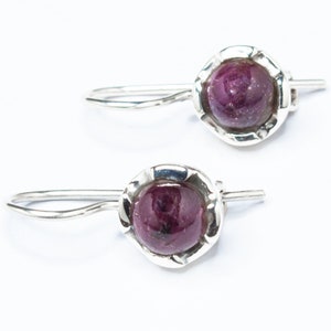 May include: A pair of silver earrings featuring a round, dark pink gemstone set in a silver bezel. The earrings have a simple, elegant design.