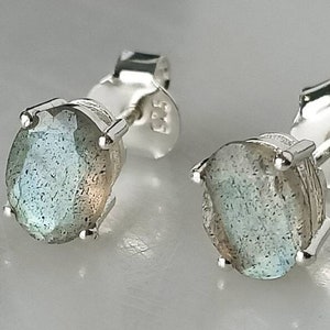 Puede incluir: Un par de pendientes de plata con piedras de labradorita ovaladas. Los pendientes tienen un acabado plateado pulido y están engastados con una sola piedra en cada pendiente.