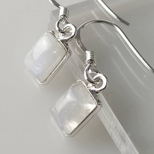 May include: Silver earrings featuring rectangular moonstone pendants. The earrings have a hook design and are set against a white backdrop. The moonstones have a milky, iridescent quality, reflecting light softly.