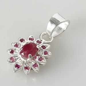 May include: A silver pendant with a flower design. The centre features a large, oval-shaped ruby, surrounded by smaller ruby accents. The pendant has a loop for a chain.