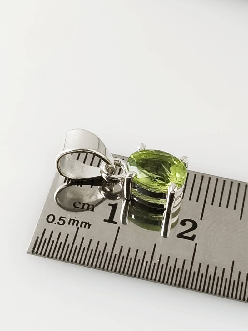 May include: A silver pendant with a green gemstone, measuring approximately 1 cm in diameter, is shown next to a ruler.