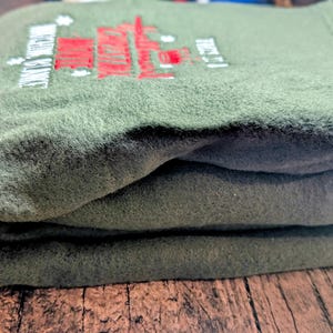 Puede incluir: Sudadera de forro polar verde oliva doblada con texto y dise&ntilde;o bordados en rojo y blanco. El texto dice "ENGINE SERVICE" y "TRUST IT". La sudadera est&aacute; apilada sobre una superficie de madera.