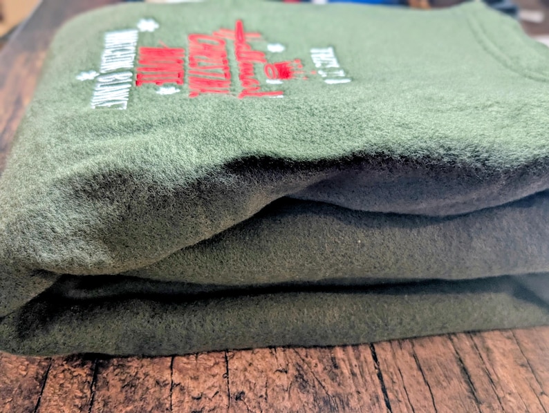 Puede incluir: Sudadera de forro polar verde oliva doblada con texto y dise&ntilde;o bordados en rojo y blanco. El texto dice "ENGINE SERVICE" y "TRUST IT". La sudadera est&aacute; apilada sobre una superficie de madera.