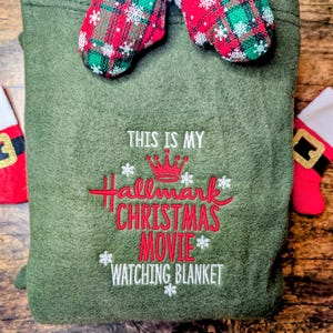 Puede incluir: Una manta verde oliva con el texto bordado en rojo "THIS IS MY Hallmark CHRISTMAS MOVIE WATCHING BLANKET". Dos mini-mitones con patrones de cuadros rojos y verdes y ribete blanco encima.