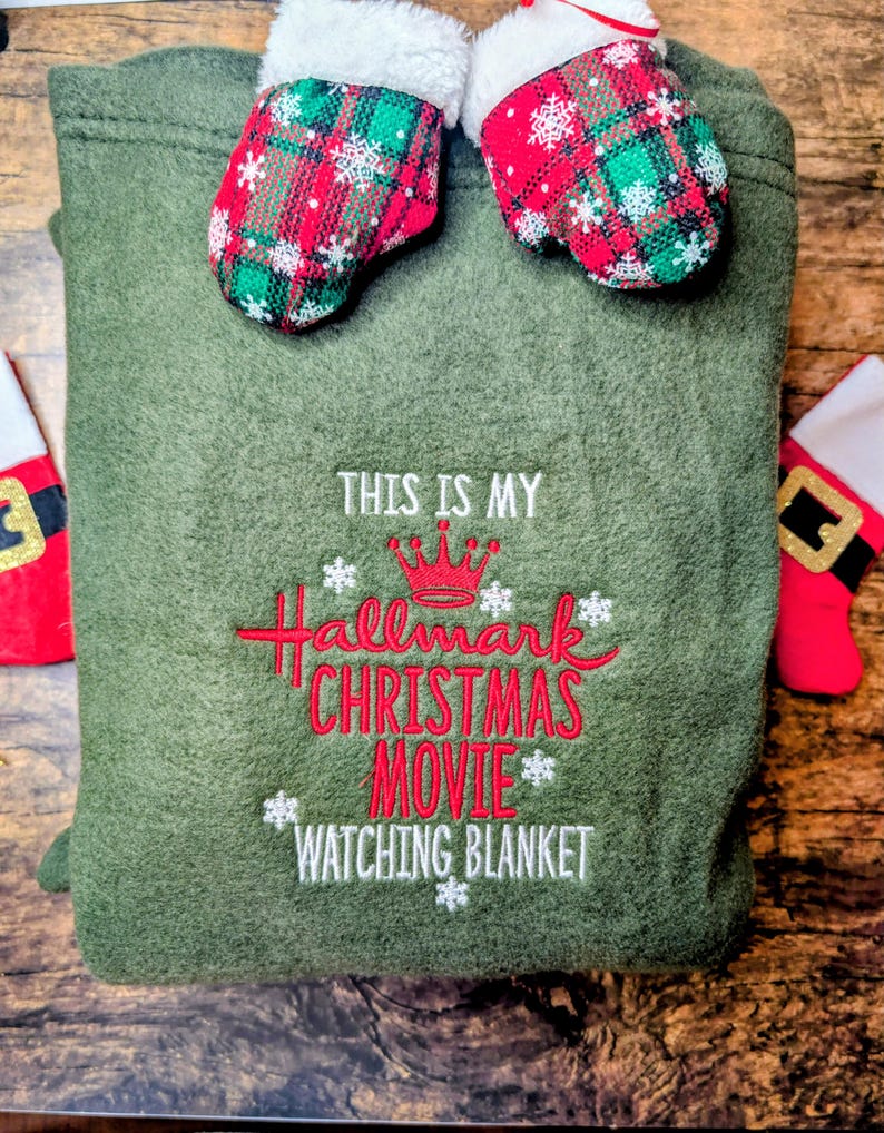 Puede incluir: Una manta verde oliva con el texto bordado en rojo "THIS IS MY Hallmark CHRISTMAS MOVIE WATCHING BLANKET". Dos mini-mitones con patrones de cuadros rojos y verdes y ribete blanco encima.