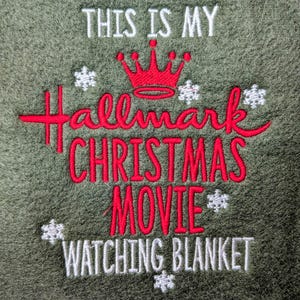 Puede incluir: Manta verde oliva bordada con la frase "THIS IS MY Hallmark CHRISTMAS MOVIE WATCHING BLANKET." El texto es en blanco y rojo, con una corona roja y acentos de copos de nieve blancos. Un art&iacute;culo tem&aacute;tico navide&ntilde;o.