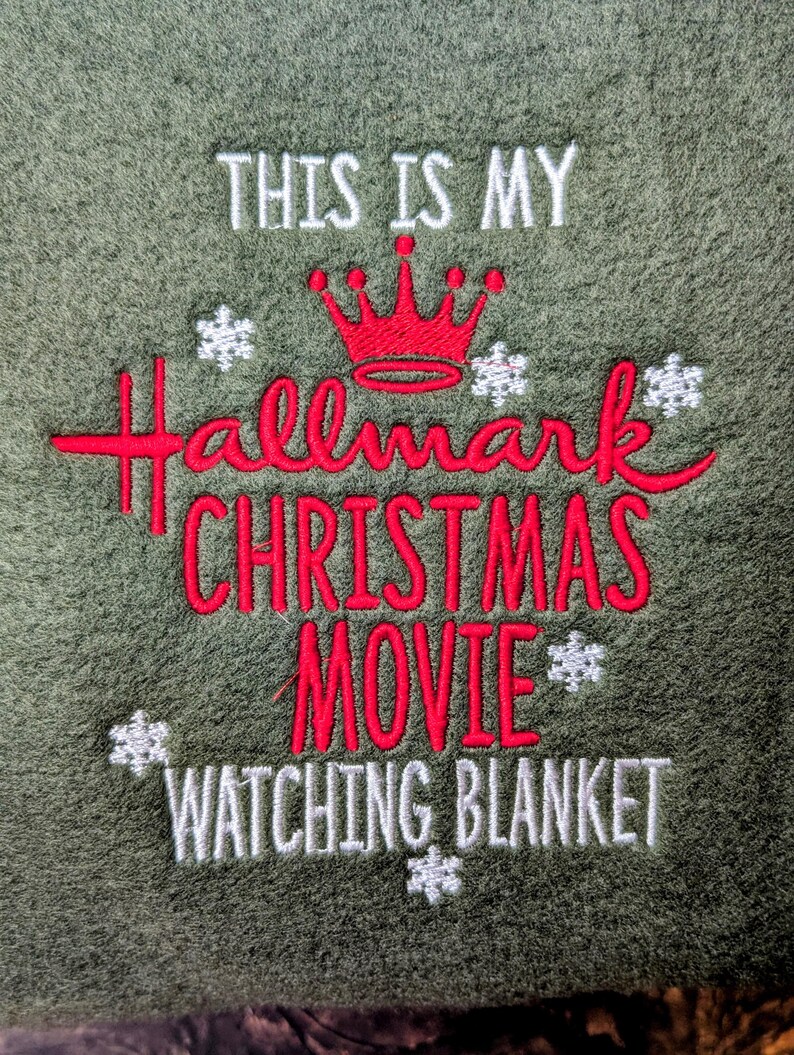 Puede incluir: Manta verde oliva bordada con la frase "THIS IS MY Hallmark CHRISTMAS MOVIE WATCHING BLANKET." El texto es en blanco y rojo, con una corona roja y acentos de copos de nieve blancos. Un art&iacute;culo tem&aacute;tico navide&ntilde;o.