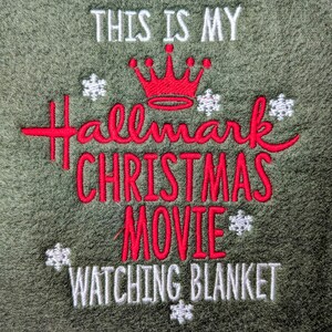 Puede incluir: Manta verde oliva con bordado: "THIS IS MY Hallmark CHRISTMAS MOVIE WATCHING BLANKET." El texto es rojo y blanco, con una corona roja y copos de nieve. Una manta acogedora para las noches de cine.