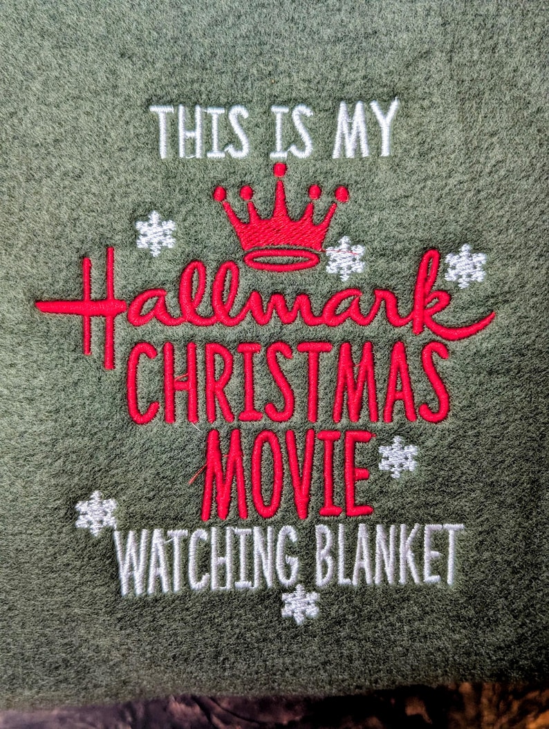 Puede incluir: Manta verde oliva con bordado: "THIS IS MY Hallmark CHRISTMAS MOVIE WATCHING BLANKET." El texto es rojo y blanco, con una corona roja y copos de nieve. Una manta acogedora para las noches de cine.
