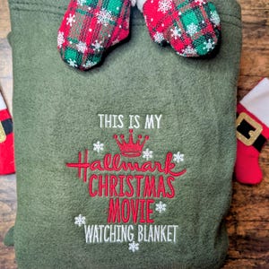 Puede incluir: Una manta verde oliva con el texto bordado "THIS IS MY Hallmark CHRISTMAS MOVIE WATCHING BLANKET" en rojo y blanco. Dos peque&ntilde;os mitones de cuadros rojos y verdes con ribete blanco descansan encima. Un art&iacute;culo festivo navide&ntilde;o.