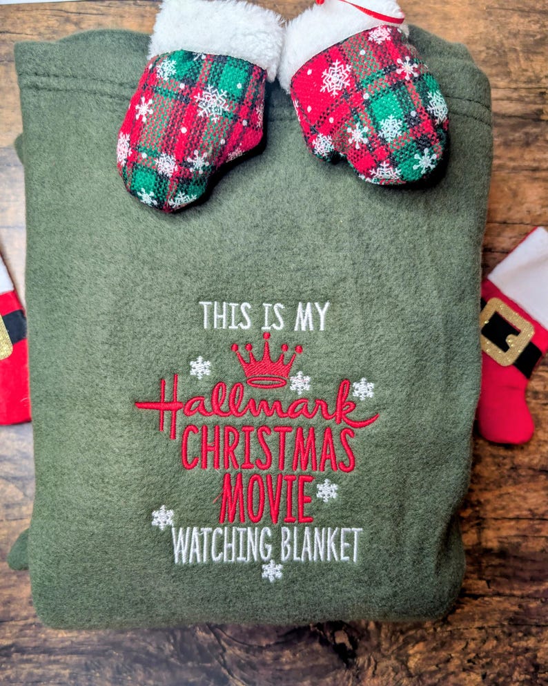 Puede incluir: Una manta verde oliva con el texto bordado "THIS IS MY Hallmark CHRISTMAS MOVIE WATCHING BLANKET" en rojo y blanco. Dos peque&ntilde;os mitones de cuadros rojos y verdes con ribete blanco descansan encima. Un art&iacute;culo festivo navide&ntilde;o.